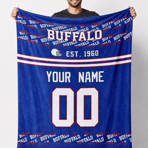 Puede incluir: Una manta azul con la palabra "BUFFALO" en blanco y el texto "EST. 1960". La manta tiene un gráfico de casco de fútbol y espacio para un nombre y un número. El número "00" se muestra en blanco con un contorno rojo.