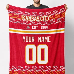 Może przedstawiać: Czerwony koc z napisem "KANSAS CITY" w białych i złotych literach. Zawiera grafikę kasku do futbolu amerykańskiego i tekst "EST. 1960". Zawiera miejsce na spersonalizowane imię i numer "00".