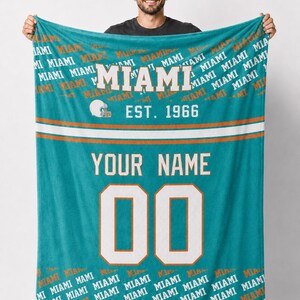 Puede incluir: Una manta turquesa con la palabra "MIAMI" en blanco y naranja, un gráfico de casco de fútbol y "EST. 1966." La manta tiene un diseño de camiseta con espacio para un nombre y el número "00."