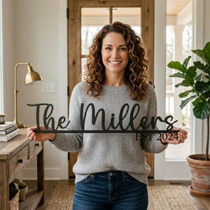 May include: A black metal sign with the words "The Millers" and "EST 2024" in a cursive font. The sign is held by a person in a gray sweater and blue jeans. The background includes a wooden door and a houseplant.