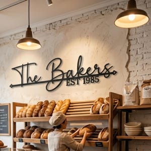 以下が含まれることがあります： 「The Bakers EST 1985」と書かれた黒い金属製の看板が、さまざまなパンのディスプレイの上にあります。パン屋の帽子をかぶった人が、木製の棚にパンを並べています。パン屋は吊り下げられたライトで照らされています。