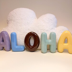 Pode incluir: Letras de cerâmica coloridas que formam a palavra "ALOHA". As letras são em tons de lavanda, azul, castanho, verde-azulado e amarelo. Um objeto branco e fofo em forma de nuvem está no fundo.
