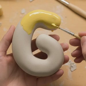 Op de afbeelding: Handen van een persoon die de witte keramische letter "G" met gele verf beschildert. De kwast brengt de verf aan op de bovenste curve van de letter. De letter wordt in de linkerhand van de persoon gehouden.