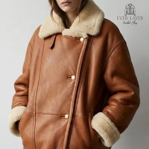Könnte beinhalten: Eine hellbraune Lederjacke mit einem cremefarbenen Lammfellkragen und passenden Lammfellbündchen. Die Jacke hat einen zweireihigen Verschluss mit goldfarbenen Knöpfen und ein Logo in der oberen rechten Ecke mit der Aufschrift EVER LAYER Jacket Shop.