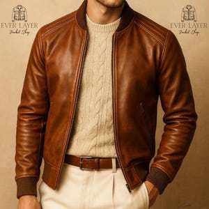Puede incluir: Una chaqueta bomber de cuero marrón con cuello y puños acanalados, sobre un suéter de punto trenzado color crema y pantalones claros con un cinturón de cuero marrón. El texto EVER LAYER Jacket Shop aparece en las esquinas superiores.