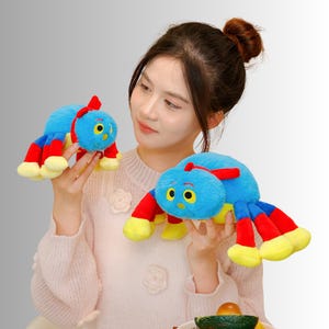 May include: Two plush caterpillar toys. The caterpillars are blue with red and yellow accents. The toys have large yellow eyes and a red antenna. The toys are being held by a person.