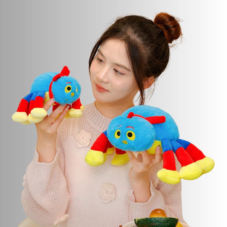 May include: Two plush caterpillar toys. The caterpillars are blue with red and yellow accents. The toys have large yellow eyes and a red antenna. The toys are being held by a person.