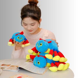 May include: Three plush toy spiders in various sizes. The spiders are blue with red and yellow legs and a red bow. One is held by a person, and two are on a magazine.