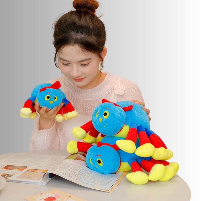 May include: Three plush toy spiders in various sizes. The spiders are blue with red and yellow legs and a red bow. One is held by a person, and two are on a magazine.