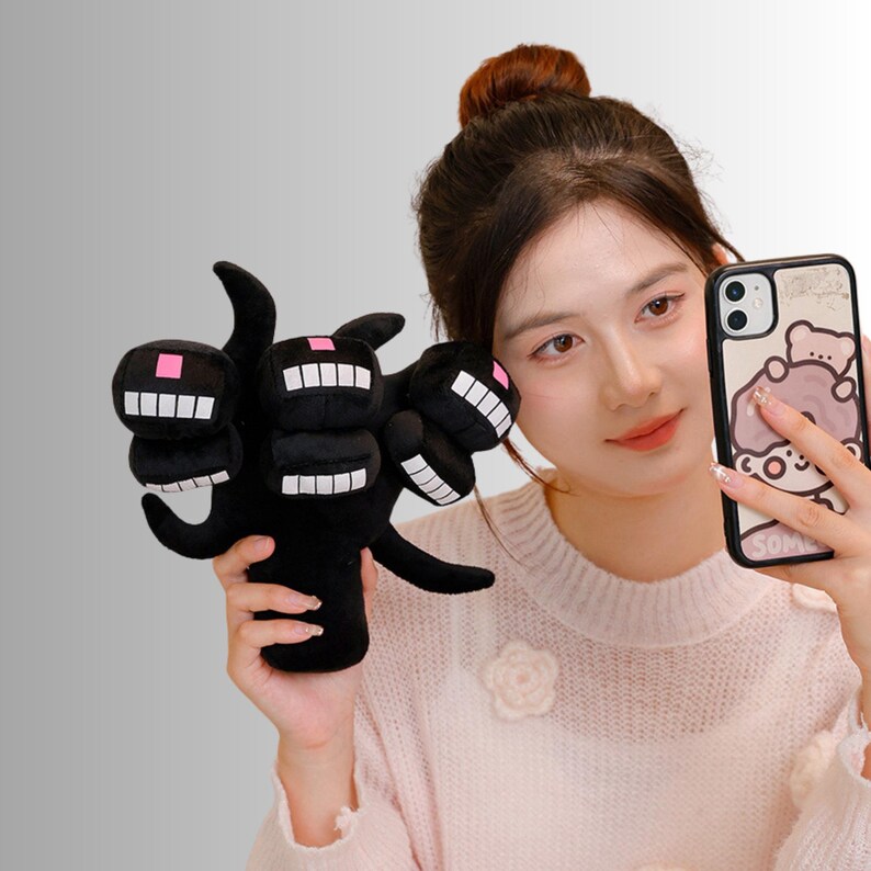 May include: Black plush toy with three heads, each with a white-toothed grin and a pink square. The toy is hand-shaped with curved fingers. The background is a light grey.
