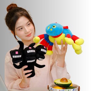 May include: Two plush toys are held in the image. One is a black, multi-headed creature with white teeth and pink squares. The other is a colourful spider-like toy with a blue head, red arms, and yellow legs. The toys appear soft and cuddly.