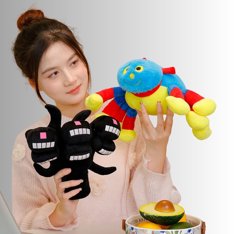 May include: Two plush toys are held in the image. One is a black, multi-headed creature with white teeth and pink squares. The other is a colourful spider-like toy with a blue head, red arms, and yellow legs. The toys appear soft and cuddly.
