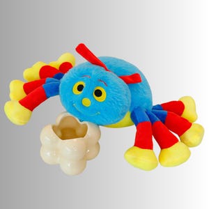 May include: A soft toy spider with a blue body, red and blue legs, and yellow details. The spider is holding a cream-coloured, cloud-shaped object. The toy has a smiling face with yellow eyes and a red antenna.