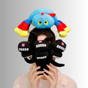 May include: A blue plush spider with red and yellow legs sits atop a person's head, while a black plush creature with multiple smiling faces is held in front. The toys are soft and colourful, perfect for children.