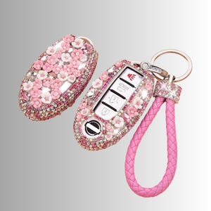 May include: A bedazzled car key fob and matching key cover. The key fob is covered in pink and white flowers and rhinestones. A pink leather strap is attached to the key fob with a silver ring.