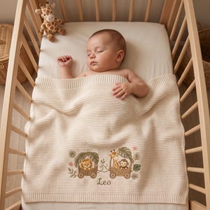 May include: A cream-coloured knit baby blanket featuring an embroidered design of two small wooden carts carrying a lion, a giraffe, and a monkey surrounded by tropical leaves. The name Leo is embroidered in brown script below the animal illustration.
