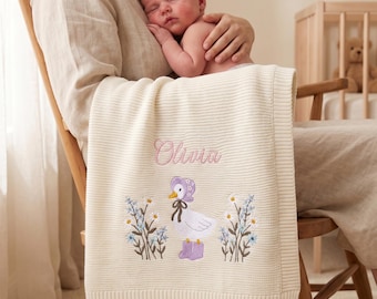 Puede incluir: Una manta de punto para bebé de color crema con un diseño bordado de un pato blanco con un gorro y botas moradas, flanqueado por flores azules y blancas. El nombre Olivia está bordado en letra cursiva rosa sobre el pato.