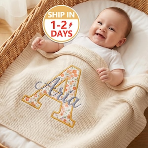May include: A cream-colored knit baby blanket featuring a large, embroidered letter A with a floral pattern in shades of orange and green. The name Aria is embroidered in a cursive grey script across the center of the letter A, which is outlined in yellow thread.