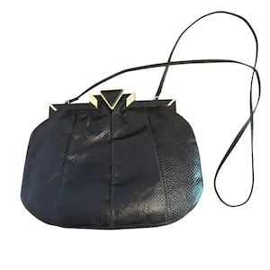 May include: A black, textured handbag with a long, thin strap. The bag has a rounded shape with a central seam and a gold-coloured clasp with a triangular accent. The strap is attached to the top of the bag.