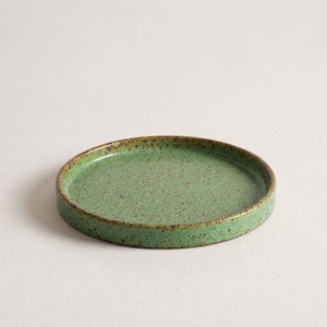 May include: A round, shallow ceramic tray featuring a speckled green glaze with brown flecks throughout. The dish has a raised edge and a smooth, flat interior surface, showcasing a consistent mottled texture across the entire piece.