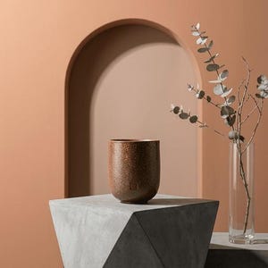 May include: A speckled brown ceramic cup with a rounded base sits on a geometric concrete block. A clear glass vase with leafy branches is to the right. The background features an archway and peach-colored walls.