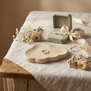 Puede incluir: Un plato decorativo beige con forma de nube que sostiene un anillo dorado, acompañado de un joyero de terciopelo verde salvia con dos pendientes, una pulsera de cadena dorada fina y pequeños ramos de flores blancas y rosas pálidas.