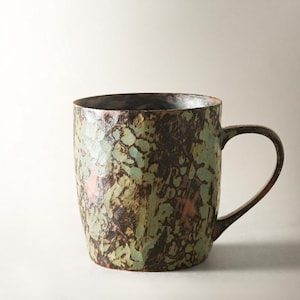 Pode incluir: Uma caneca de cerâmica com uma superfície texturizada e manchada, apresentando uma mistura de tons castanho-escuro, verde-sálvia e toques de cobre. O recipiente tem um corpo cilíndrico com base ligeiramente afunilada e uma asa curva.