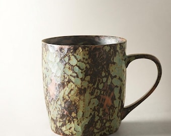 May include: A ceramic mug featuring a textured, mottled surface with a blend of dark brown, sage green, and hints of copper tones. The vessel has a cylindrical body with a slightly tapered base and a curved handle on the right side.