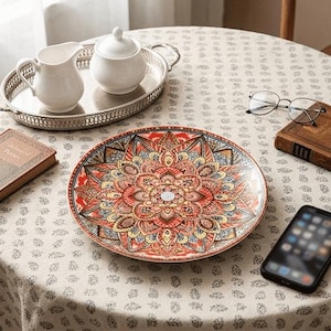 Op de afbeelding: Een decoratieve keramische plaat met een gedetailleerd bloemenmandala-ontwerp in rood, oranje, geel en blauw. De plaat staat op een tafel met een zilveren dienblad, een melkkan, een suikerpot en een smartphone.