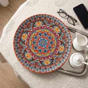 Puede incluir: Un plato decorativo de cerámica con un diseño de mandala intrincado y vibrante en rojo, azul, amarillo y naranja. El plato tiene un borde rojo y está colocado sobre un mantel estampado. Una bandeja con una jarra de crema y un azucarero está al lado del plato.