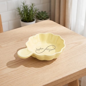 May include: A pale yellow ceramic dish with a scalloped edge and a small handle. The centre of the dish features a dark grey infinity symbol graphic with the names Sophia and James printed in a cursive script font inside the bowl.