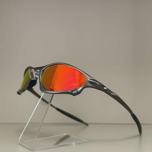 May include: Sport sunglasses with a dark gray frame and vibrant red lenses. The sleek design features a curved shape and is displayed on a clear acrylic stand. The lenses have a reflective quality.