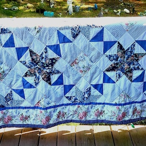May include: A rectangular quilt featuring a patchwork design of blue and white star patterns, light blue gingham squares, and floral fabric sections with pink roses. The border displays a continuous floral print with pink roses on a white background.