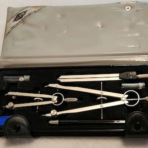 May include: An Alvin precision drawing kit in a rectangular case with a grey lid featuring the brand logo. The black interior tray holds several silver metal drafting compasses, a black handle tool, and two small clear plastic lead containers.
