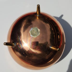 May include: A polished copper bowl with a smooth, reflective surface and three tapered brass legs. The bowl features a small, circular, light-colored sticker with an indistinct emblem centered on the underside of the rounded base.