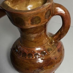May include: A brown glazed ceramic pitcher with a rounded body, a narrow neck, and a curved handle. The surface features a glossy finish with darker brown circular indentations and decorative dotted patterns around the neck and midsection.