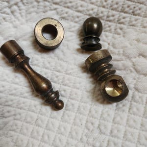 May include: A collection of four brass-toned metal hardware components, including a slender finial, a small circular washer, a spherical knob, and a threaded cylindrical piece with a side opening, all featuring a weathered, aged metallic finish.