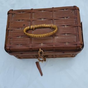 May include: A rectangular brown woven basket with a flat lid and a single oval-shaped handle attached to the top. The basket features a grid-like weave pattern and includes a small, dangling brown tag secured by a thin cord at the front center.