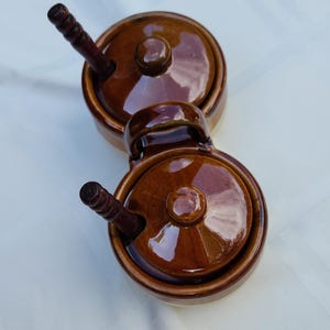 Puede incluir: Un juego de dos tarros para condimentos de cerámica esmaltada en color marrón, unidos entre sí, con tapas a juego y cucharas de madera oscura. Cada tarro cuenta con una base circular, una tapa con pomo central y un asa lateral.