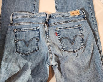 May include: Light blue denim jeans featuring a classic five-pocket design, a tan leather patch on the back waistband, and a small red tab on the right back pocket. The jeans have a bootcut leg silhouette with visible stitching details.