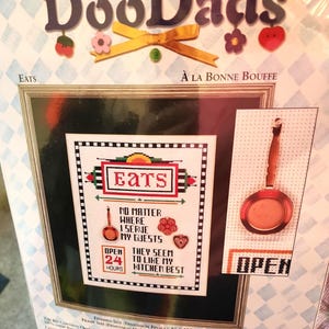 Op de afbeelding: Een Needle Treasures DooDads kruissteekpakket met een keukenthema, inclusief de tekst EATS en NO MATTER WHERE I SERVE MY GUESTS THEY SEEM TO LIKE MY KITCHEN BEST. De set bevat een afbeelding van een koekenpan en een afmeting van 20 cm bij 25 cm.