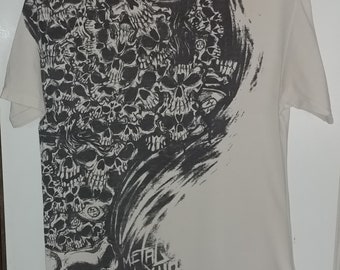 May include: Off-white t-shirt featuring a large graphic of skulls in shades of gray and black. The design covers the front and part of the right side, with the words "METAL MULISHA" printed at the bottom. The shirt has short sleeves and a crew neck.
