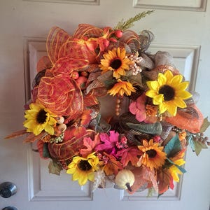 May include: A decorative wreath featuring yellow sunflowers, orange and red mesh ribbon, artificial autumn leaves in shades of red and orange, small white pumpkins, and clusters of berries. The arrangement is circular and dense with textures.