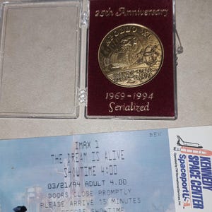 Może przedstawiać: Złota moneta pamiątkowa z okazji 25. rocznicy Apollo XI w przezroczystym etui, z napisem "KENNEDY SPACE CENTER FLORIDA" i "1969-1994". Poniżej niebieski bilet z napisem "THE DREAM IS ALIVE" i "SHOWTIME 4:00".