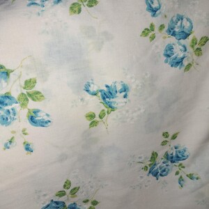 May include: White fabric with a pattern of blue roses and green leaves. The roses are in various stages of bloom, scattered across the surface. The fabric has a soft, slightly blurred appearance.