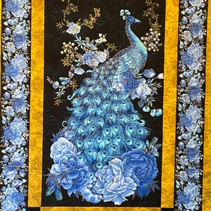 May include: A blue and gold peacock with its tail feathers spread out, surrounded by blue and white flowers on a black background. The peacock is standing on a bed of blue and white flowers. The background is a black fabric with a gold border.
