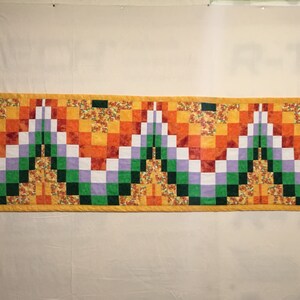May include: A colorful quilt with a geometric pattern in shades of orange, yellow, green, white, and purple. The quilt is made up of squares and rectangles, and the pattern is symmetrical.