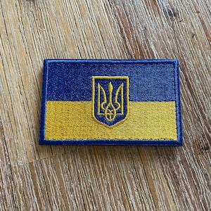 May include: Embroidered patch featuring the Ukrainian flag. The design includes a blue upper half and a yellow lower half, with a gold trident emblem in the center. The patch has a blue border and is rectangular in shape.