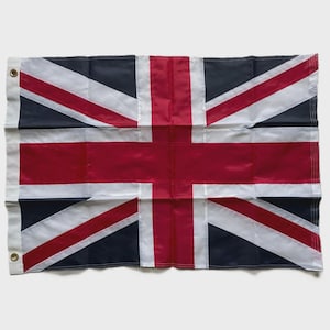 May include: A Union Jack flag, with a design of red, white, and navy blue. The flag has a white border and metal eyelets for hanging. The red and white cross and diagonal stripes are prominent.