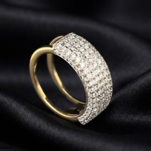 May include: A gold ring featuring a wide, curved band section encrusted with multiple rows of small, clear, sparkling stones. The band transitions into a smooth, polished gold finish on the sides, creating a distinct contrast between the textured, stone-set surface and the solid metal structure of the ring.